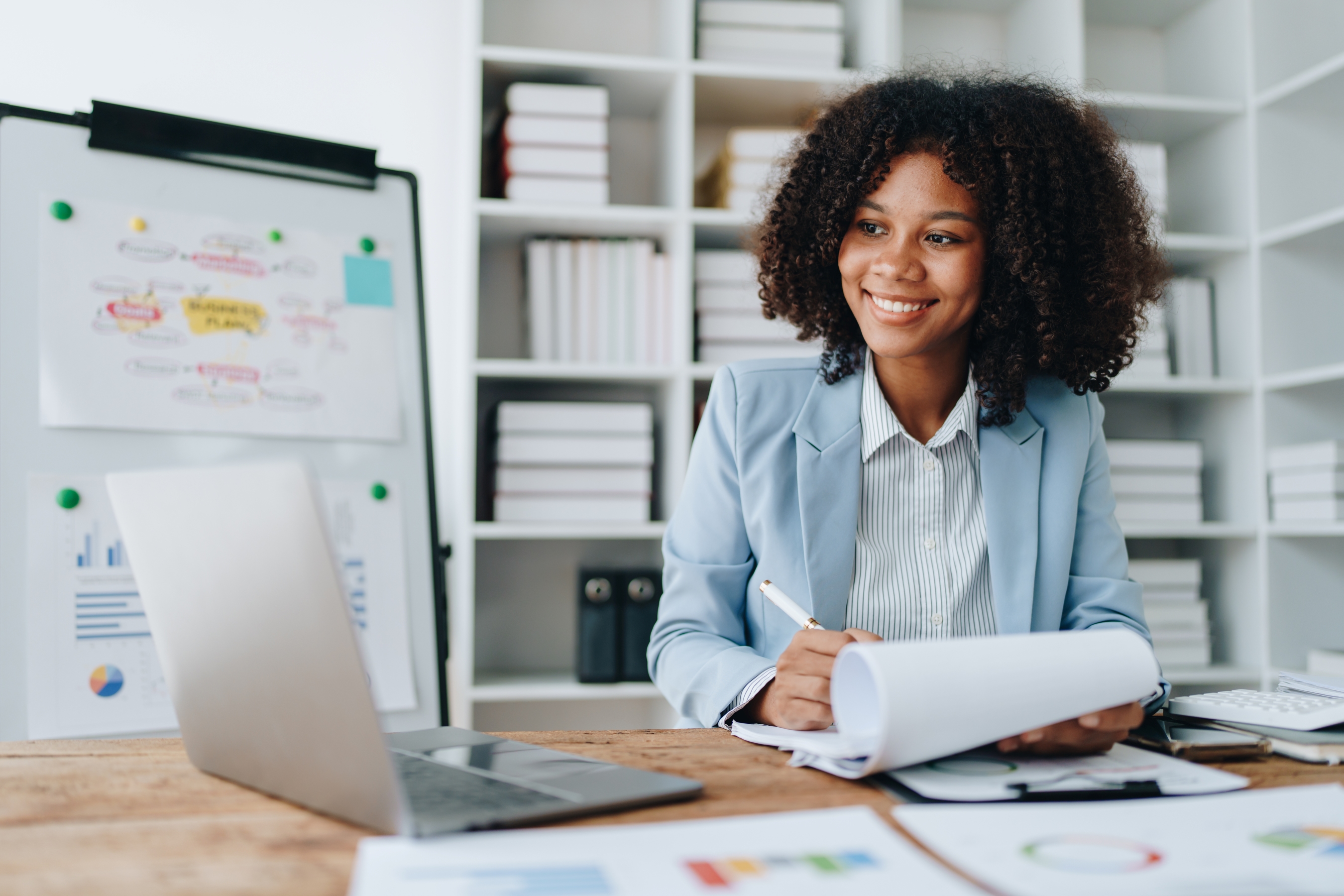 Woman with papers and a laptop at a desk in front of a white board.